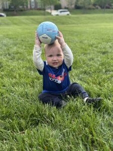 A young toddler sitting in a soccer field holding a soccer ball above his head
